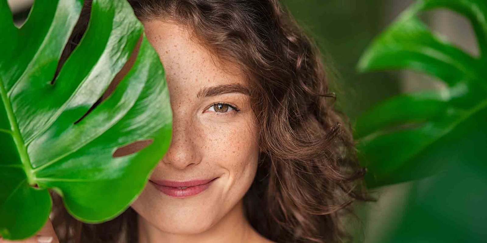 Close up face of beautiful young woman covering her face by green monstera leaf while looking at camera. Natural smiling girl with green palm leaf. Portrait of beauty woman with natural makeup and freckles on skin standing behind big green leaves.